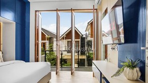 Una habitación de hotel moderna con un amplio ventanal corredero que da a un balcón, una cama, un televisor y una mesita con una planta.