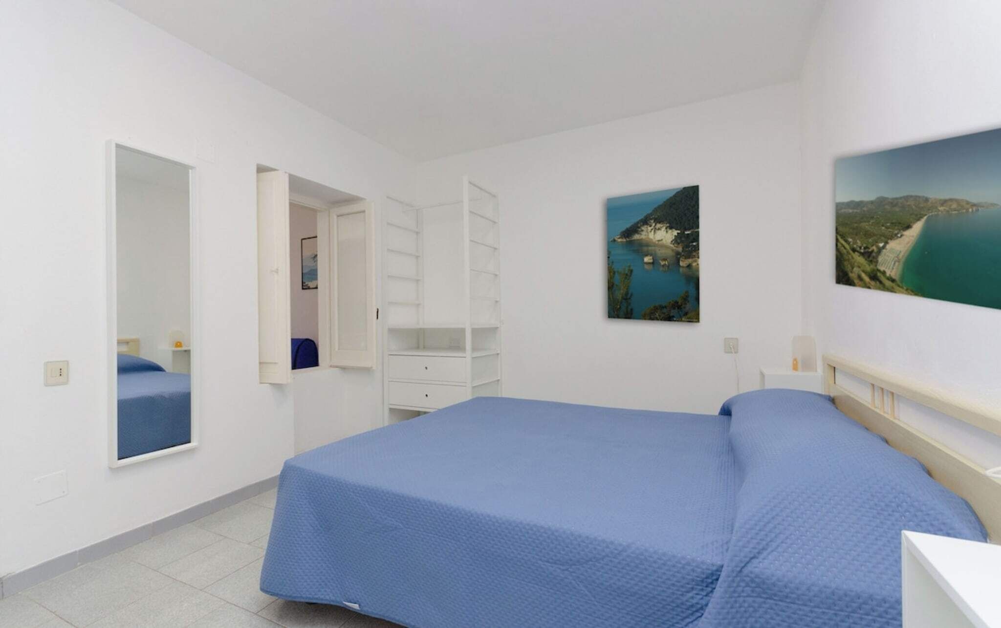 Quarto com cama, guarda-roupa, espelho e um quadro de uma paisagem costeira.