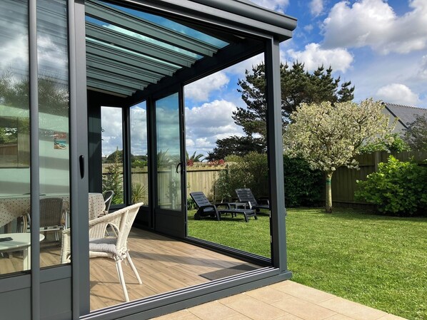 Un patio moderno con puertas de vidrio, mobiliario de mimbre y vistas a un jardín con césped y árboles.