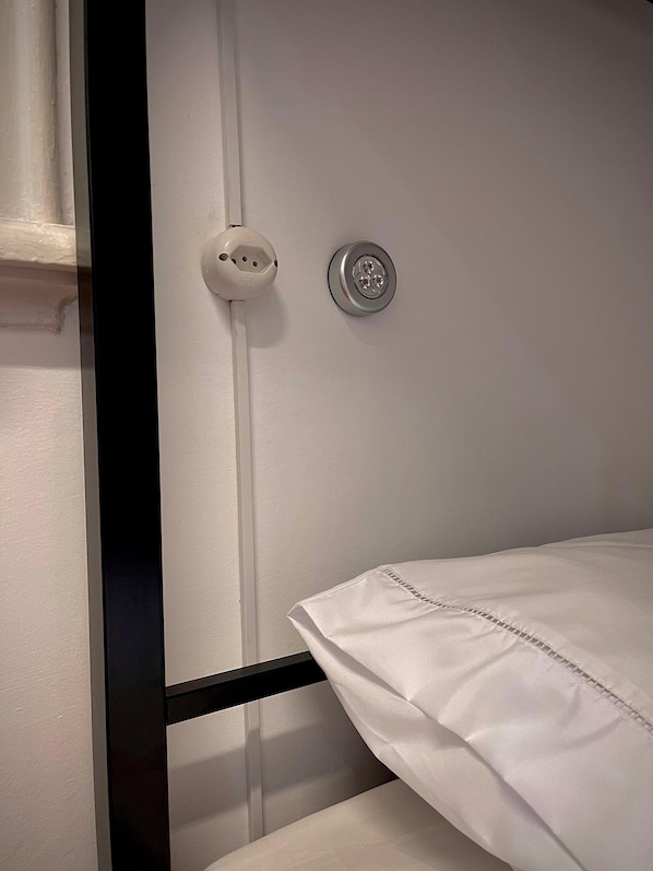 A bunk bed with a white mattress and a metal frame. There is a wall socket and a light switch on the wall. - Urban Nomad Hostel & Food (São Paulo)