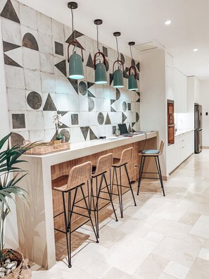 A modern kitchen with a geometric backsplash, pendant lights, a central island with bar stools, and a refrigerator. - Casa du Soleil Hostal Boutique (Madrid)