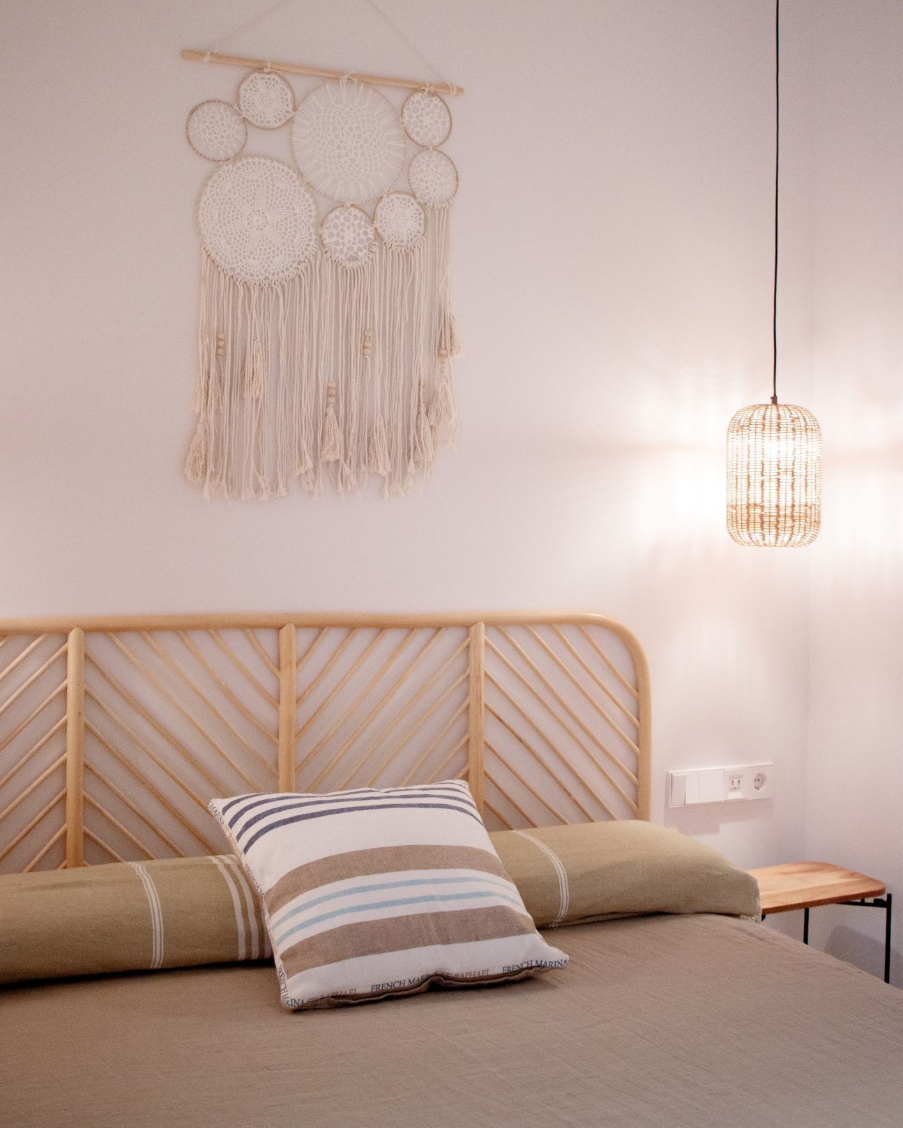 Un dormitorio con cabecera de madera, cama con almohadas a rayas, un tapiz de macramé y una lámpara colgante.