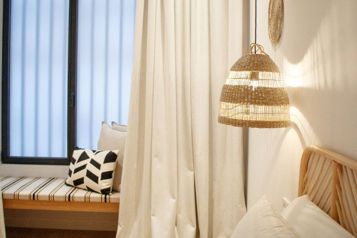 A bedroom with a bed, a window with blinds, a bedside table, and a hanging light fixture.