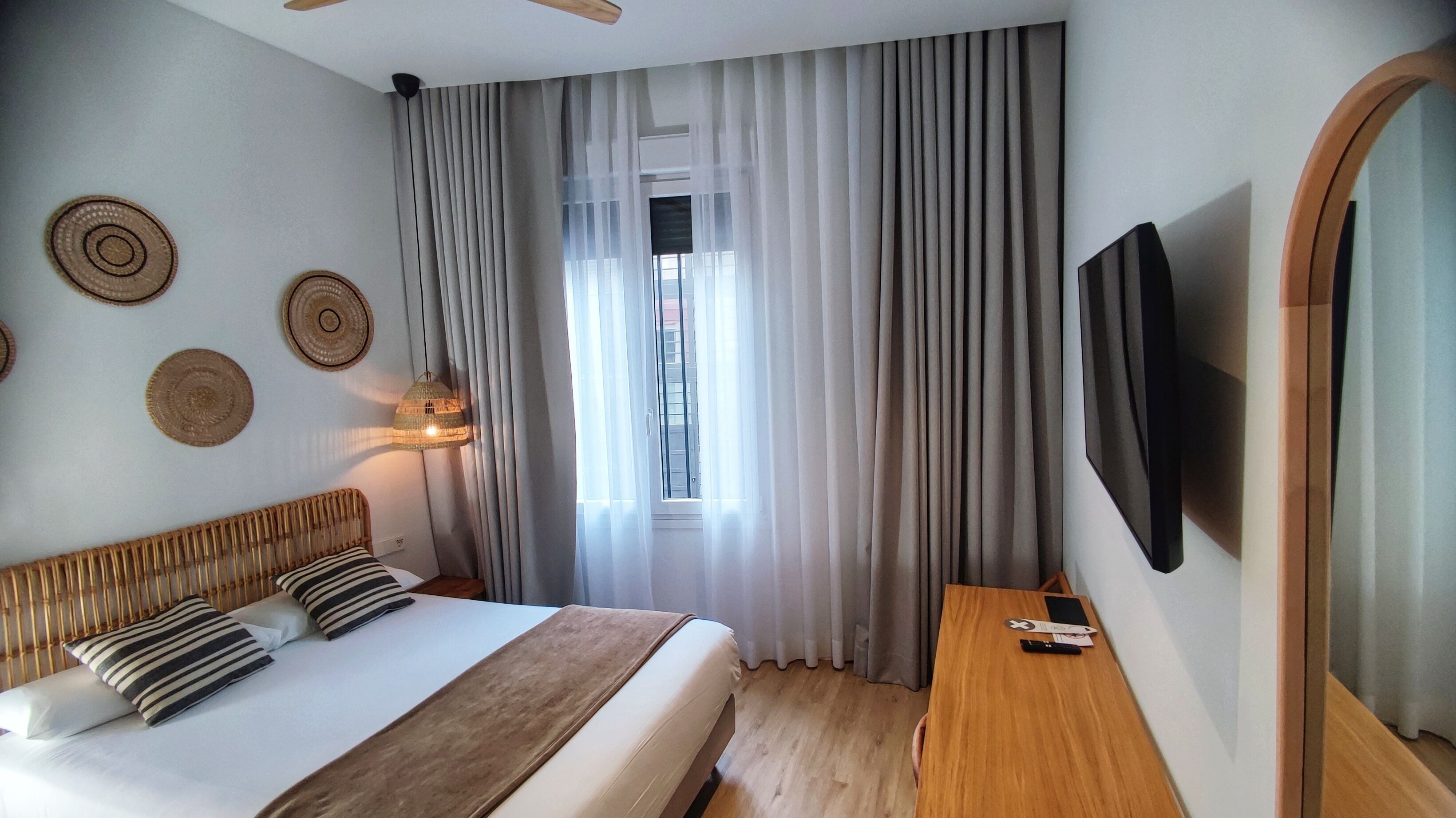 Habitación de hotel moderna con una cama grande, cabecera de madera, televisor de pantalla plana, mesita de noche de madera y una ventana con cortinas translúcidas.