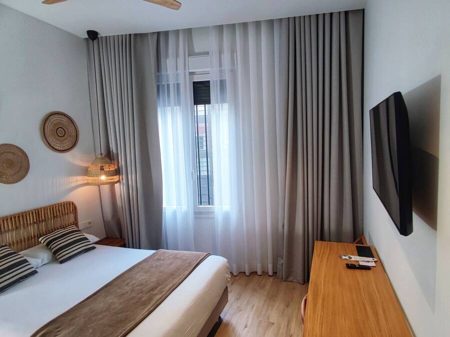 Habitación de hotel moderna con una cama grande, cabecera de madera, televisor de pantalla plana, mesita de noche de madera y una ventana con cortinas translúcidas.