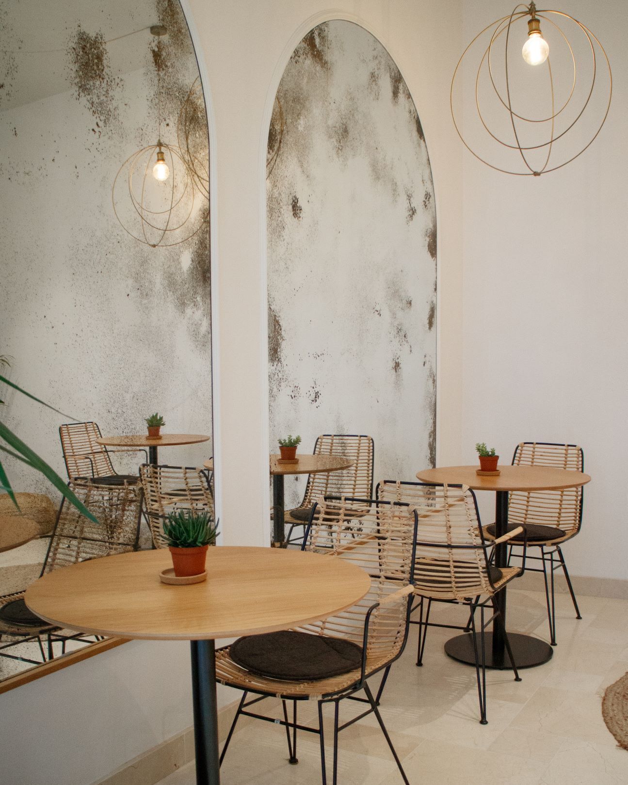 Un interior rústico con mesas de madera redondas, sillas tejidas, un espejo grande y una lámpara colgante.