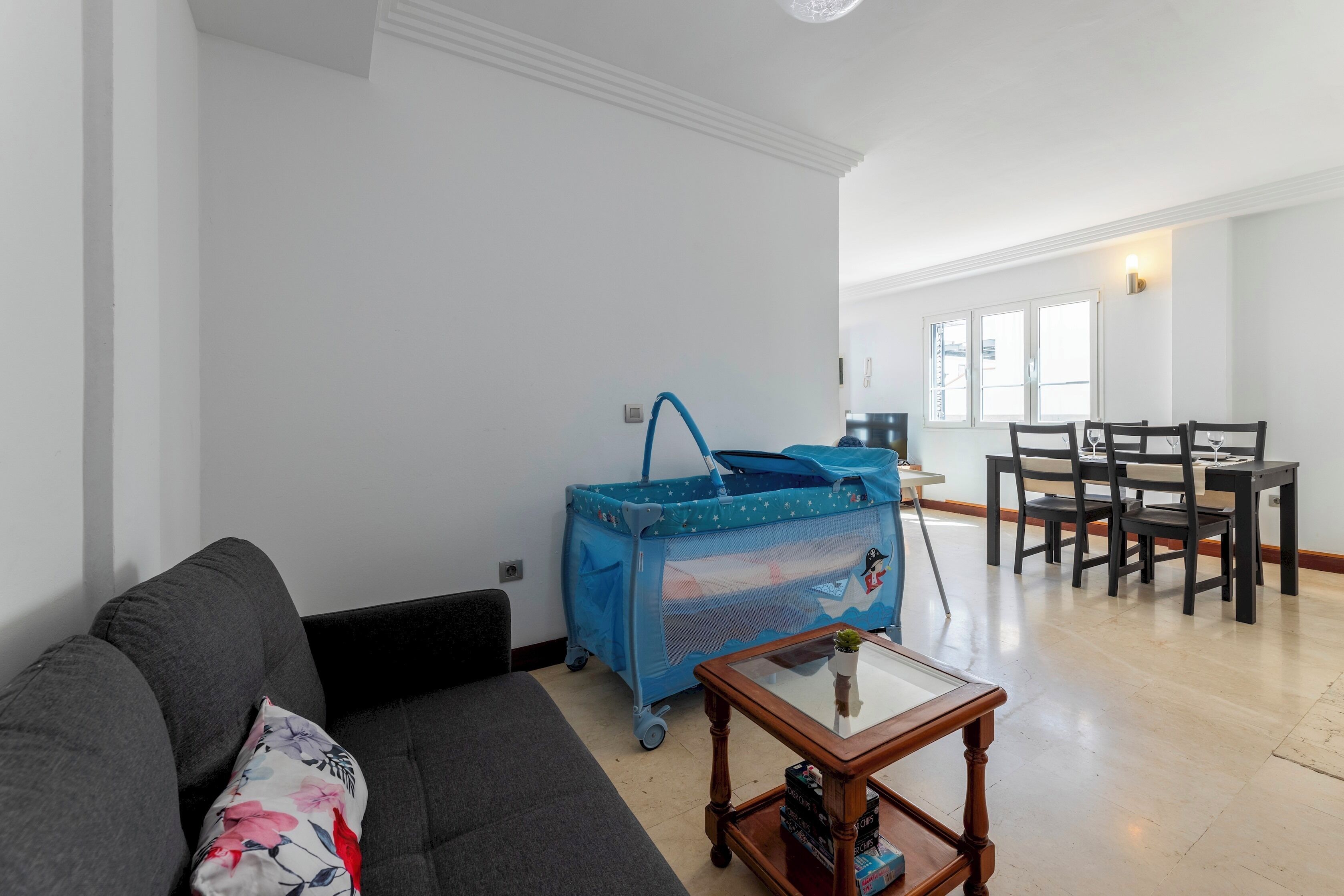 A living room with a grey sofa, a coffee table, a blue baby stroller, and a dining area with chairs and a table.