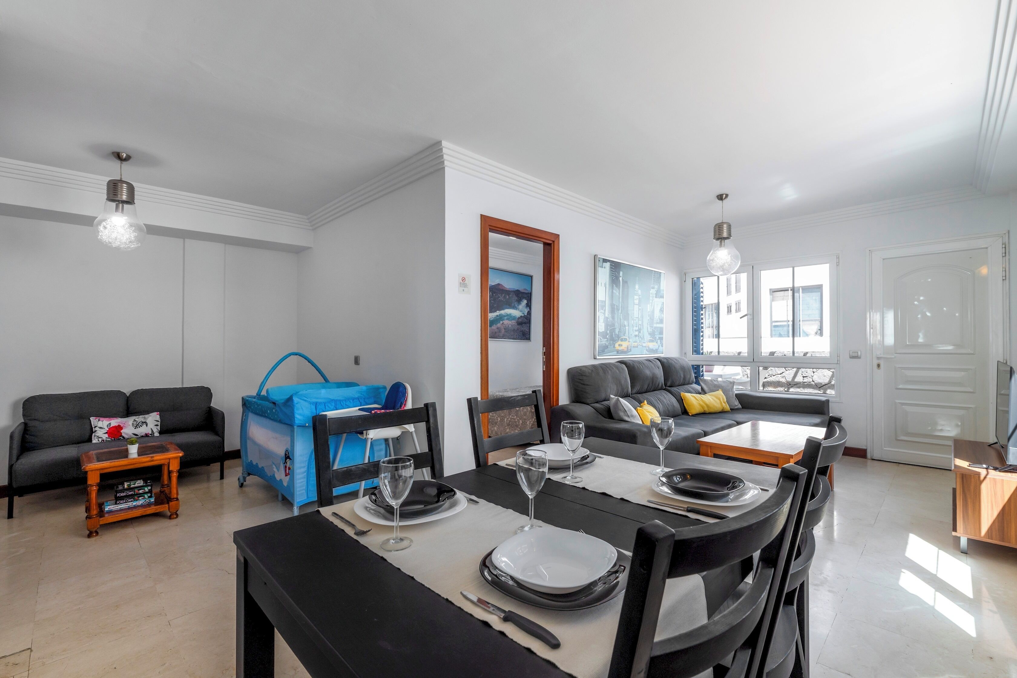A modern living room with a dining area, grey sofas, a blue playpen, and a large window.
