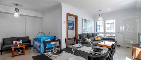 A modern living room with a dining area, grey sofas, a blue playpen, and a large window.