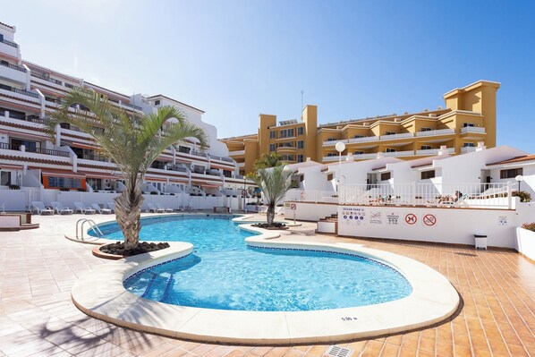 A swimming pool surrounded by a white building with balconies, palm trees, and a clear blue sky.