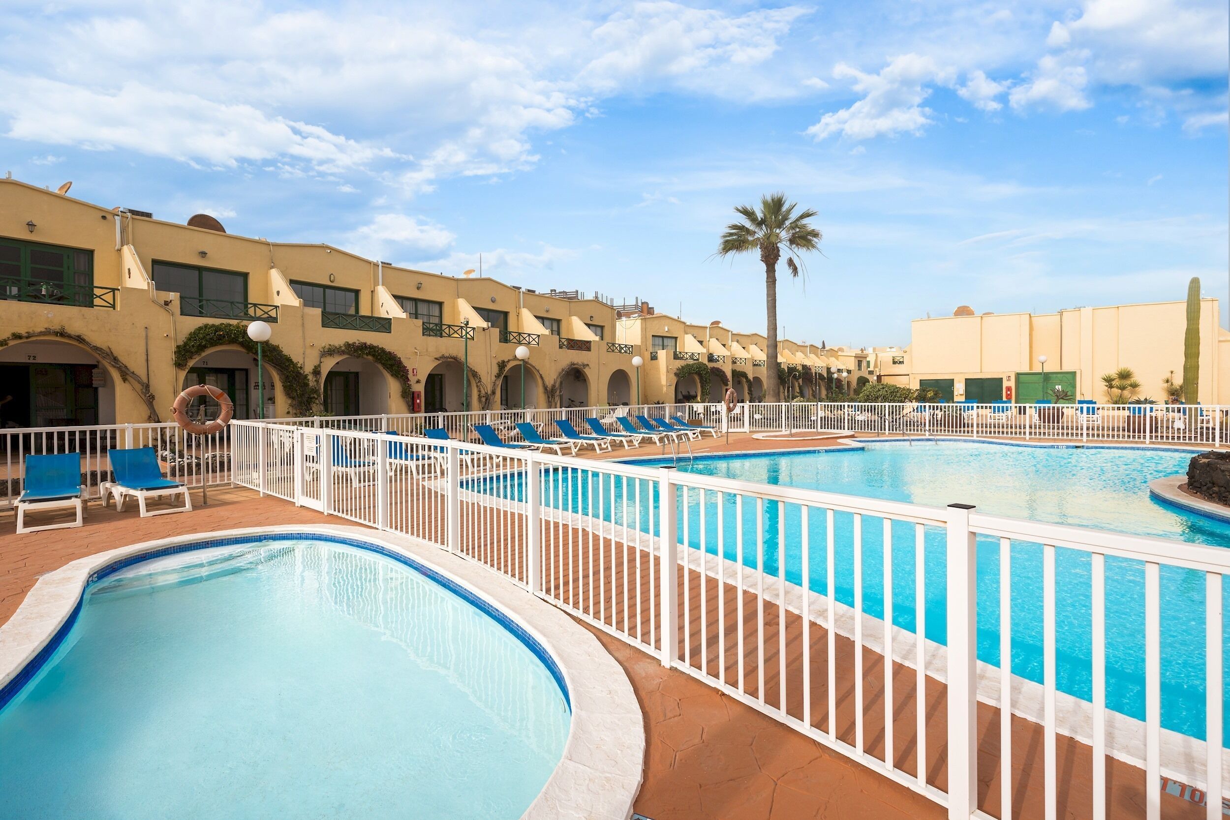 A swimming pool area with multiple pools and lounge chairs, surrounded by a white fence.