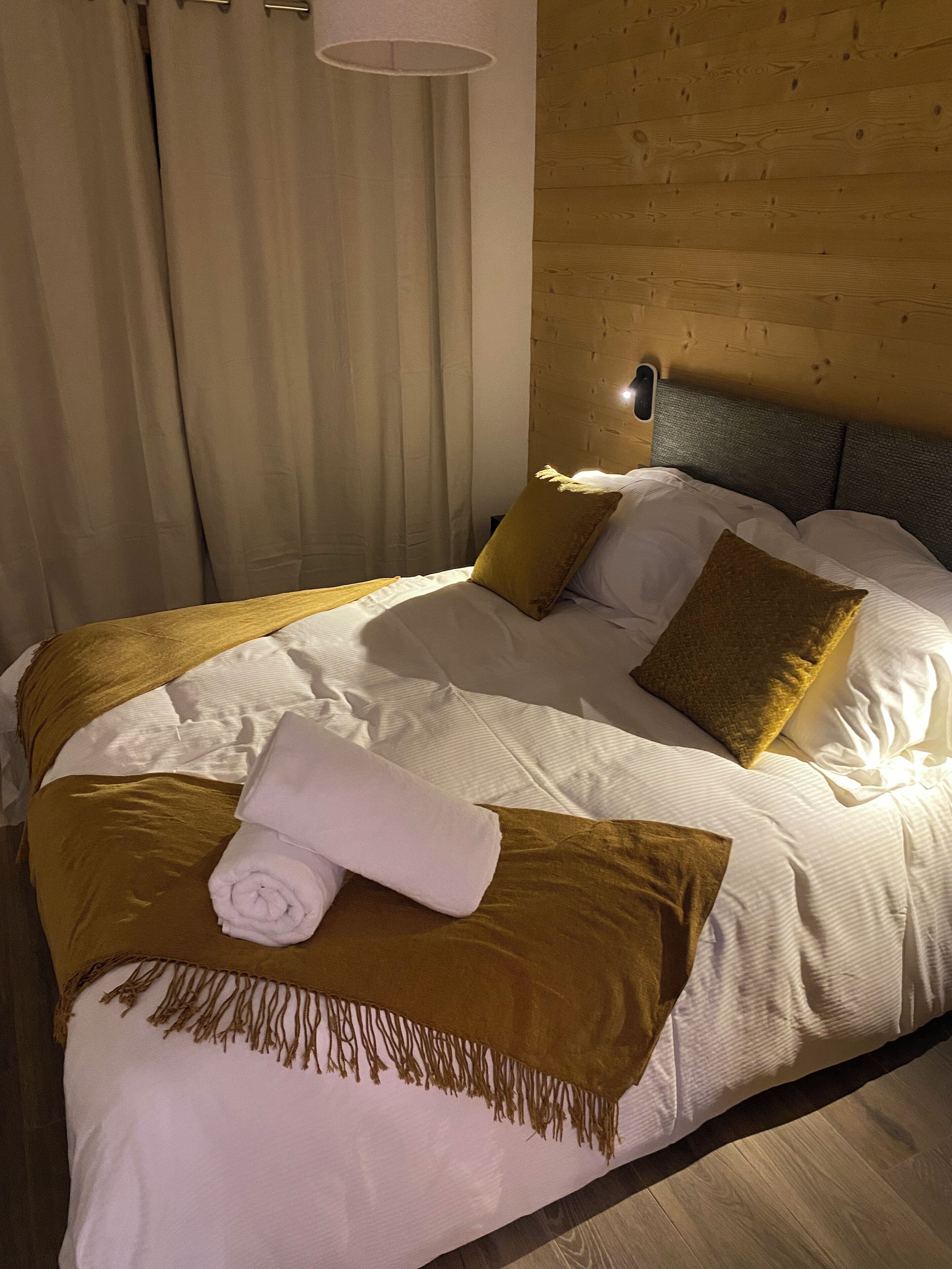 Un lit bien fait, avec une literie blanche et marron, une serviette enroulée et une tête de lit dotée d’un luminaire.