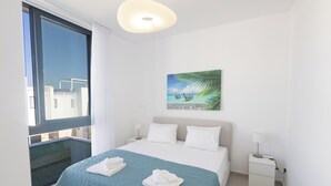 A hotel room with a large bed, a nightstand, a painting of a beach scene, and a view of buildings through a window.