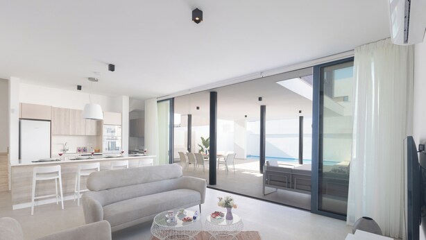 A modern living room with a sofa, coffee table, and TV. A kitchen is visible in the background with a dining area and large windows.