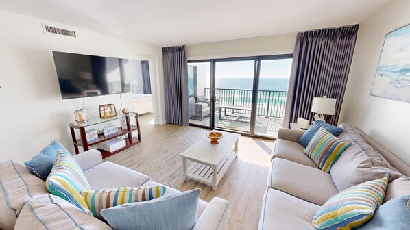 A modern living room with a sofa set, a coffee table, and a flat-screen TV. There is a balcony with a view of the ocean.