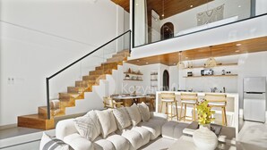 Una sala moderna con un sofá blanco grande, un comedor con sillas de madera, una cocina con armarios blancos y una escalera de madera.