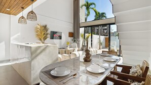 Un comedor moderno con una gran mesa ovalada dispuesta para cuatro personas, un televisor montado en la pared y vistas a palmeras a través de amplios ventanales.