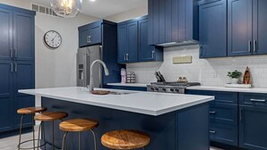 A kitchen with dark blue cabinets, a white island with stools, stainless steel appliances, and a clock on the wall. - Juniper Presents: Beach Bliss Home with Rooftop Deck & Pool Access (Union Pier)