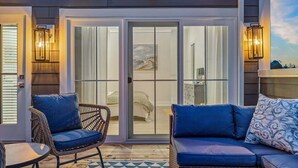 A covered outdoor area with a sliding glass door leading to a bedroom, wicker furniture, and wall-mounted lights. - Juniper Presents: Beach Bliss Home with Rooftop Deck & Pool Access (Union Pier)
