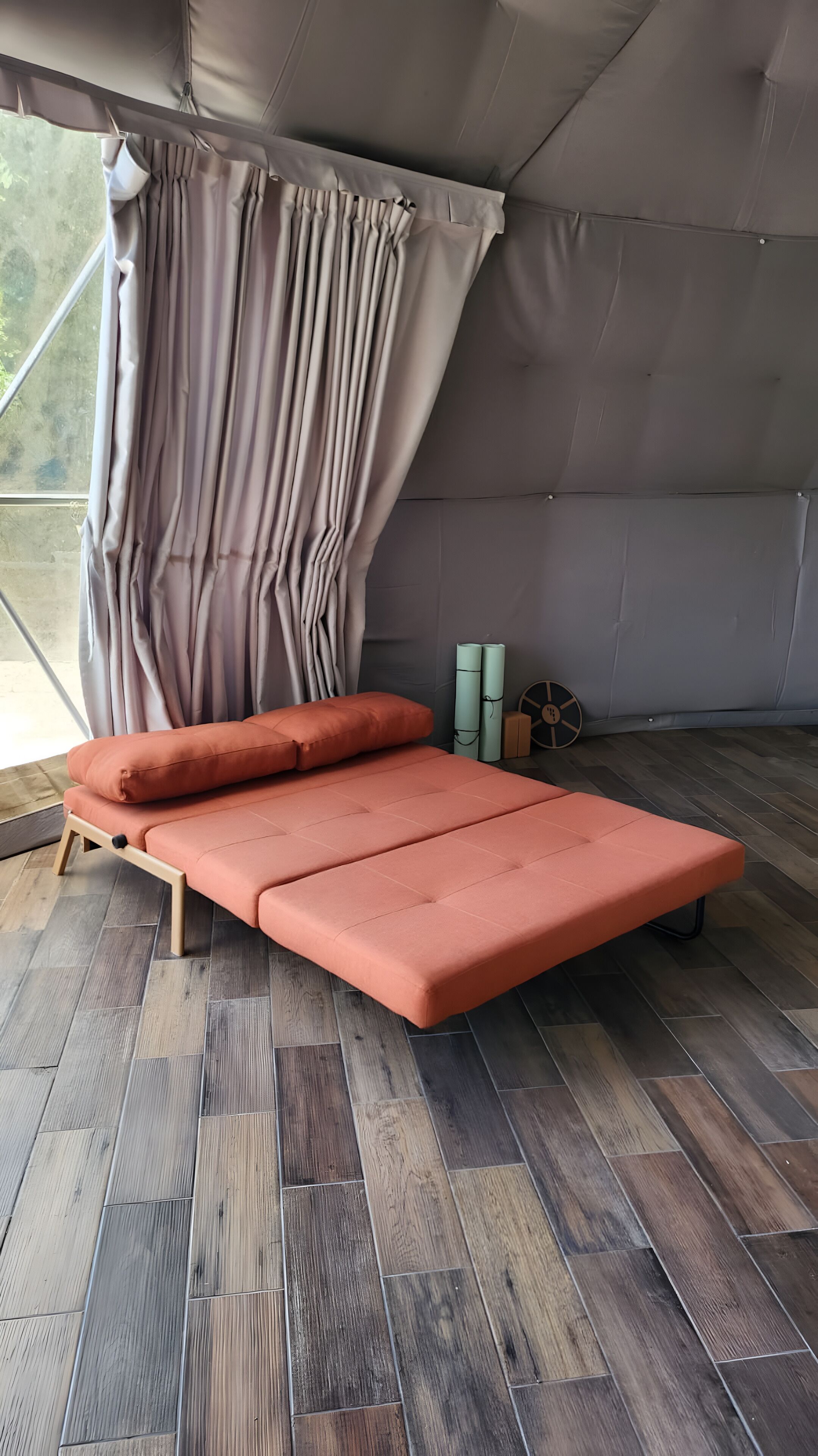 A single bed with a wooden frame and a mattress, a window with curtains, and a view of the outdoors.