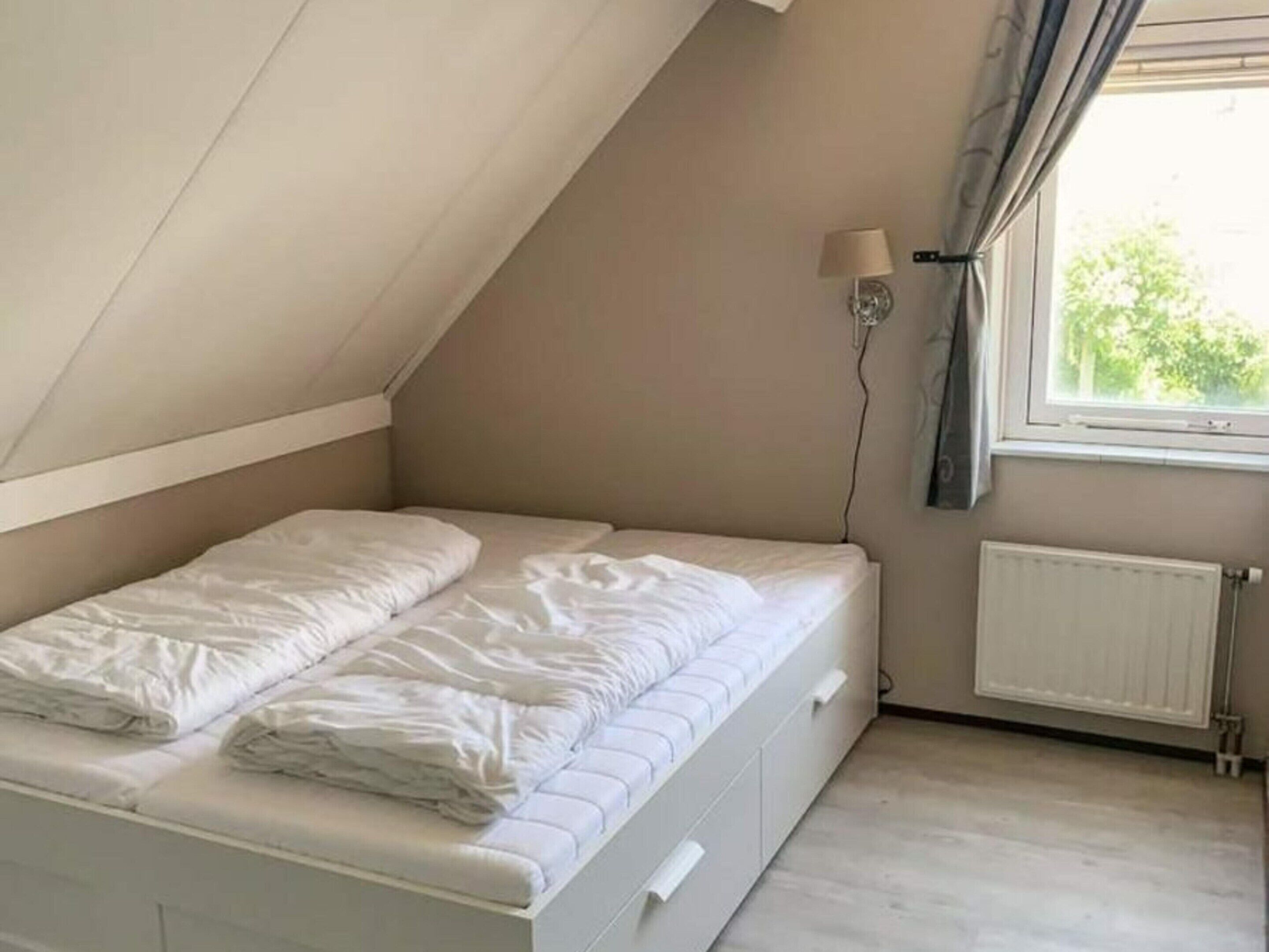 Une petite chambre avec un lit simple, un plafond en pente, une fenêtre avec des rideaux, une lampe fixée au mur et un radiateur.
