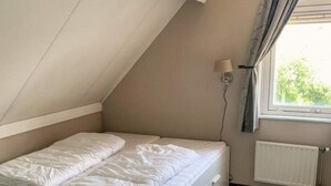 Una habitación pequeña con cama individual, techo inclinado, ventana con cortinas, lámpara fijada a la pared y radiador.
