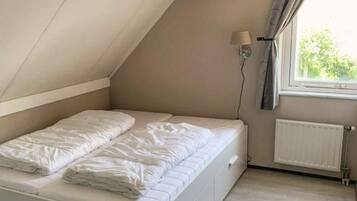 Une petite chambre avec un lit simple, un plafond en pente, une fenêtre avec des rideaux, une lampe fixée au mur et un radiateur.