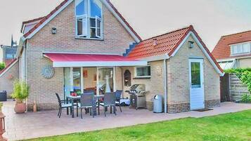 Une maison à deux étages avec un toit recouvert de tuiles rouges, une véranda à l’avant avec une table et des chaises, et un petit garage attenant.