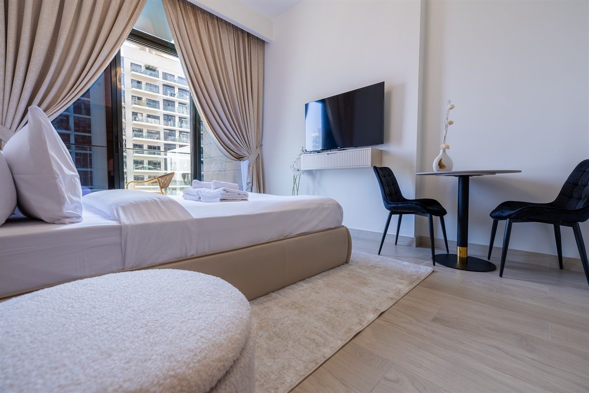 A hotel room with a bed, a TV, a small table, two chairs, and a view of a building outside.