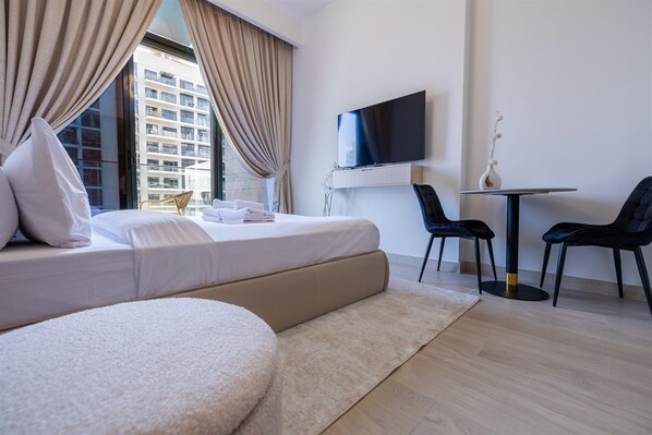A hotel room with a bed, a TV, a small table, two chairs, and a view of a building outside.