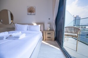 A hotel room with a bed, a nightstand, a chair, a window with a view of buildings, and a wall-mounted mirror.
