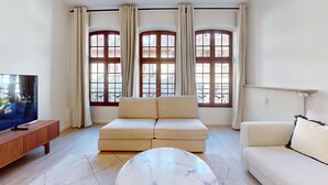 A living room with a sofa, a coffee table, a TV on a wooden stand, and large windows with curtains. - Hallebardes Cathédrale (Strasbourg)