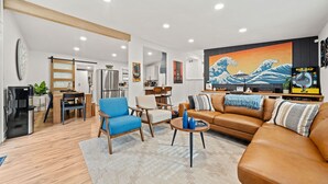 Una sala de estar moderna con un sofá de cuero marrón, un sillón azul y una mesa de centro. Se observa una zona de cocina con refrigerador, estufa y mesa de comedor. Un cuadro grande de una ola adorna la pared.