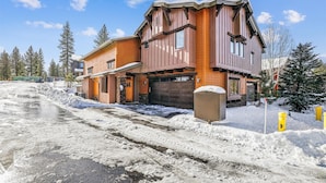 A two-story wooden house with a garage, surrounded by snow and trees.