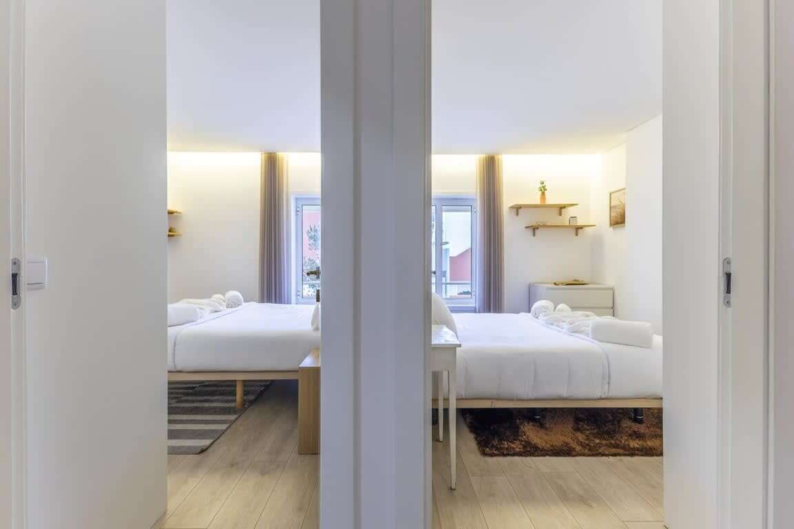 A hotel room with two beds, a bedside table, a nightstand, a window with curtains, and a view of the outside.