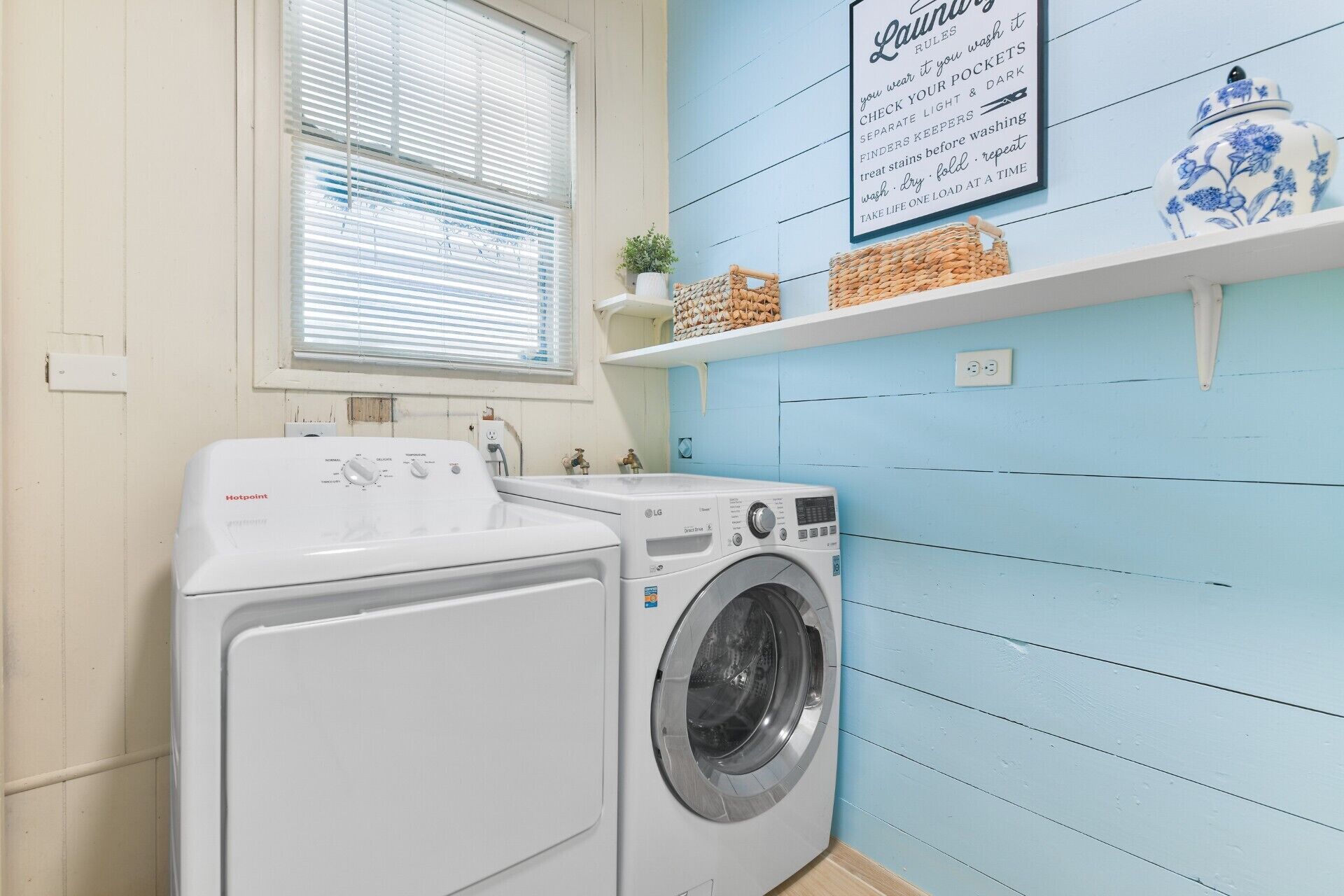 A laundry area with a washing machine and dryer, a window with blinds, a shelf with baskets, and a laundry rules sign.