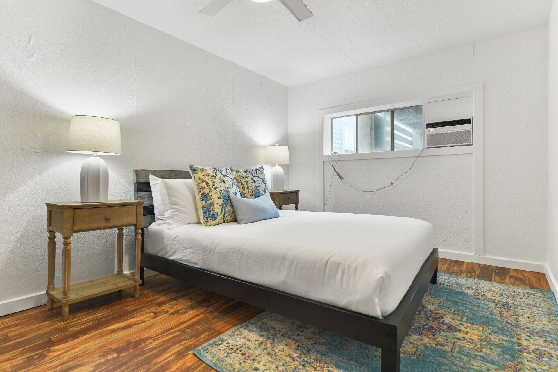 A bedroom with a bed, wooden nightstands, a window with an air conditioning unit, and a patterned rug.