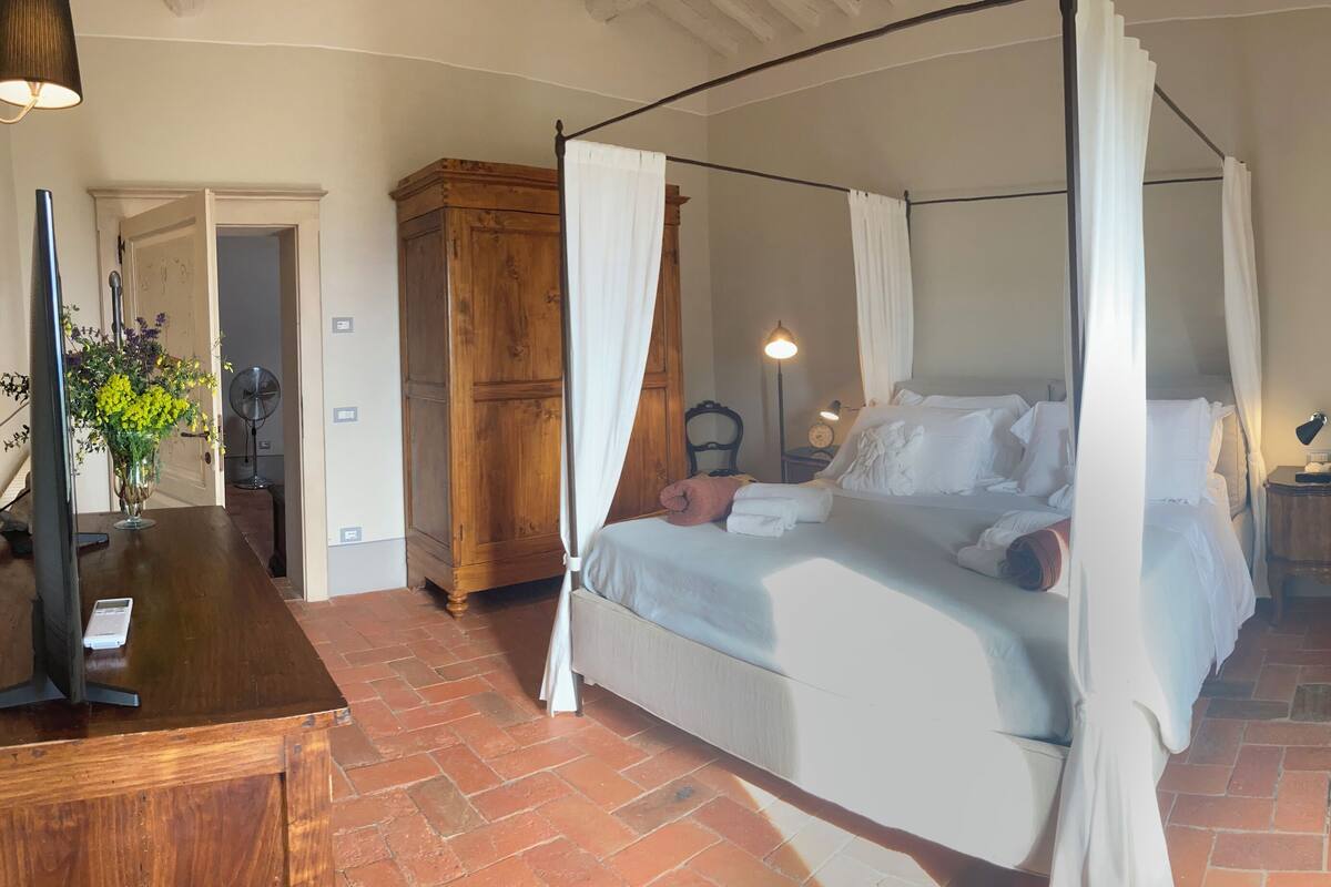 Un lit à baldaquin à quatre poteaux, avec une table de chevet en bois et un bureau équipé d’un ordinateur et agrémenté d’un vase de fleurs.