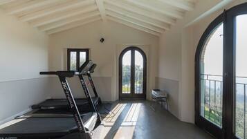 Une pièce spacieuse avec un plafond haut, un tapis de course, une porte donnant sur un balcon et un projecteur posé sur un support.
