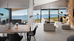 Una sala moderna con un ventanal grande que ofrece vistas panorámicas, un comedor con mesa y sillas de madera, y una zona de estar acogedora con sofá y reposapiés.