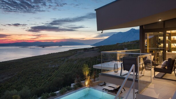 Un espacio exterior moderno con piscina, asientos y vistas a un cuerpo de agua y montañas al atardecer.