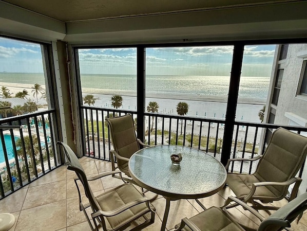 Ein Balkon mit einem Glastisch und vier Stühlen, der Blick auf einen Strand und den Ozean gewährt.