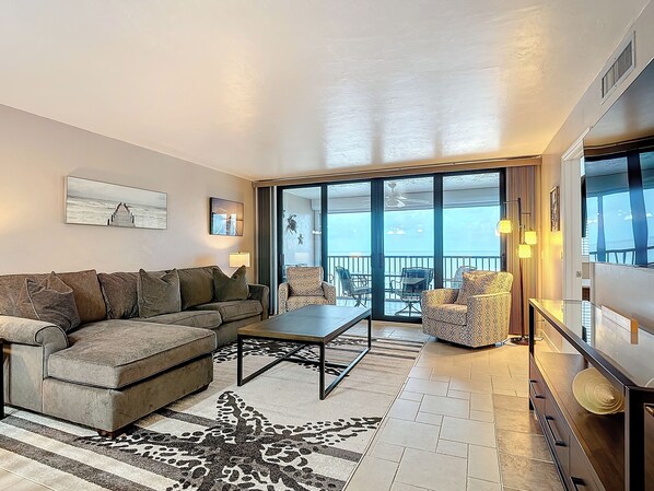 A modern living room with a sofa, coffee table, and patterned rug. It features a large sliding glass door leading to a balcony with outdoor seating.