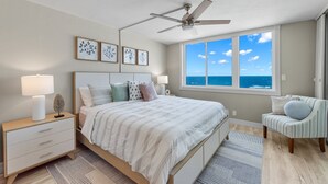 Ein Schlafzimmer mit Bett, Nachttisch, Lampe, Stuhl und Blick auf das Meer durch ein Fenster.