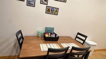 Comedor con mesa de madera, cuatro sillas, individuales, cesta de aperitivos y cuadros en la pared.