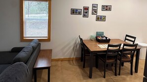 Un espacio habitable compacto con zona de comedor, sofá, ventana con persianas e imágenes enmarcadas en la pared.