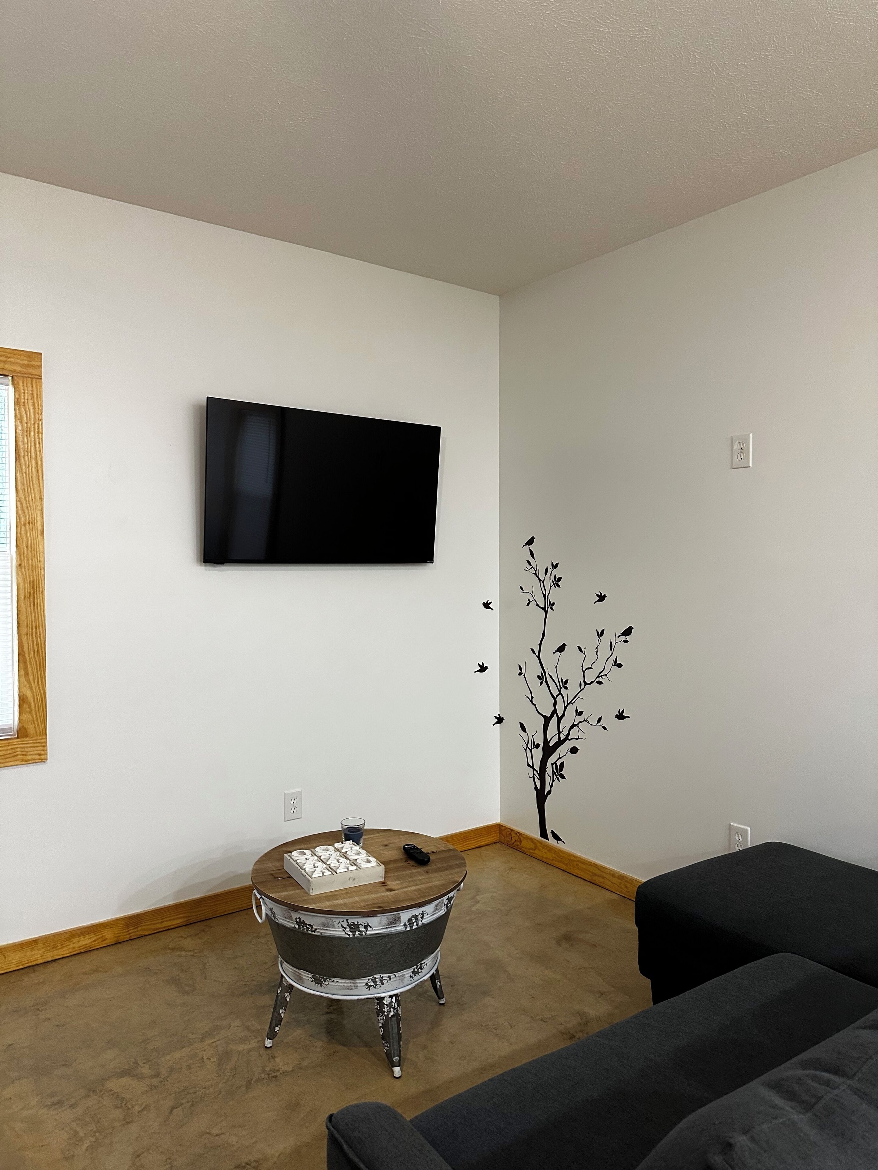 Una sala de estar con un televisor de pantalla plana montado en la pared, una mesa redonda de madera con un vaso, y un sofá negro.