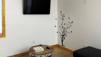 Una sala de estar con un televisor de pantalla plana montado en la pared, una mesa redonda de madera con un vaso, y un sofá negro.