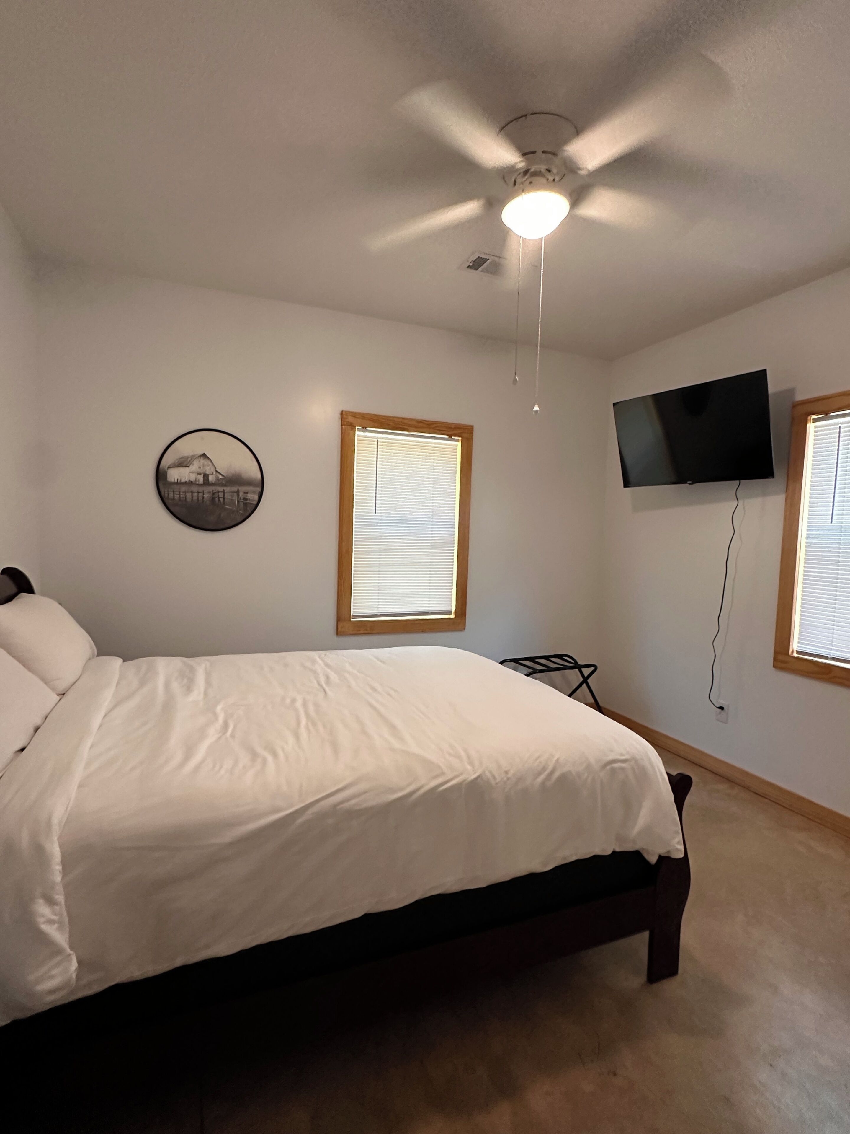 Un dormitorio con cama, ventilador de techo, televisor montado en la pared y ventana con persianas.