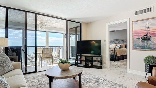 A spacious living area with a large TV, comfortable seating, a balcony with a view, and a painting on the wall.